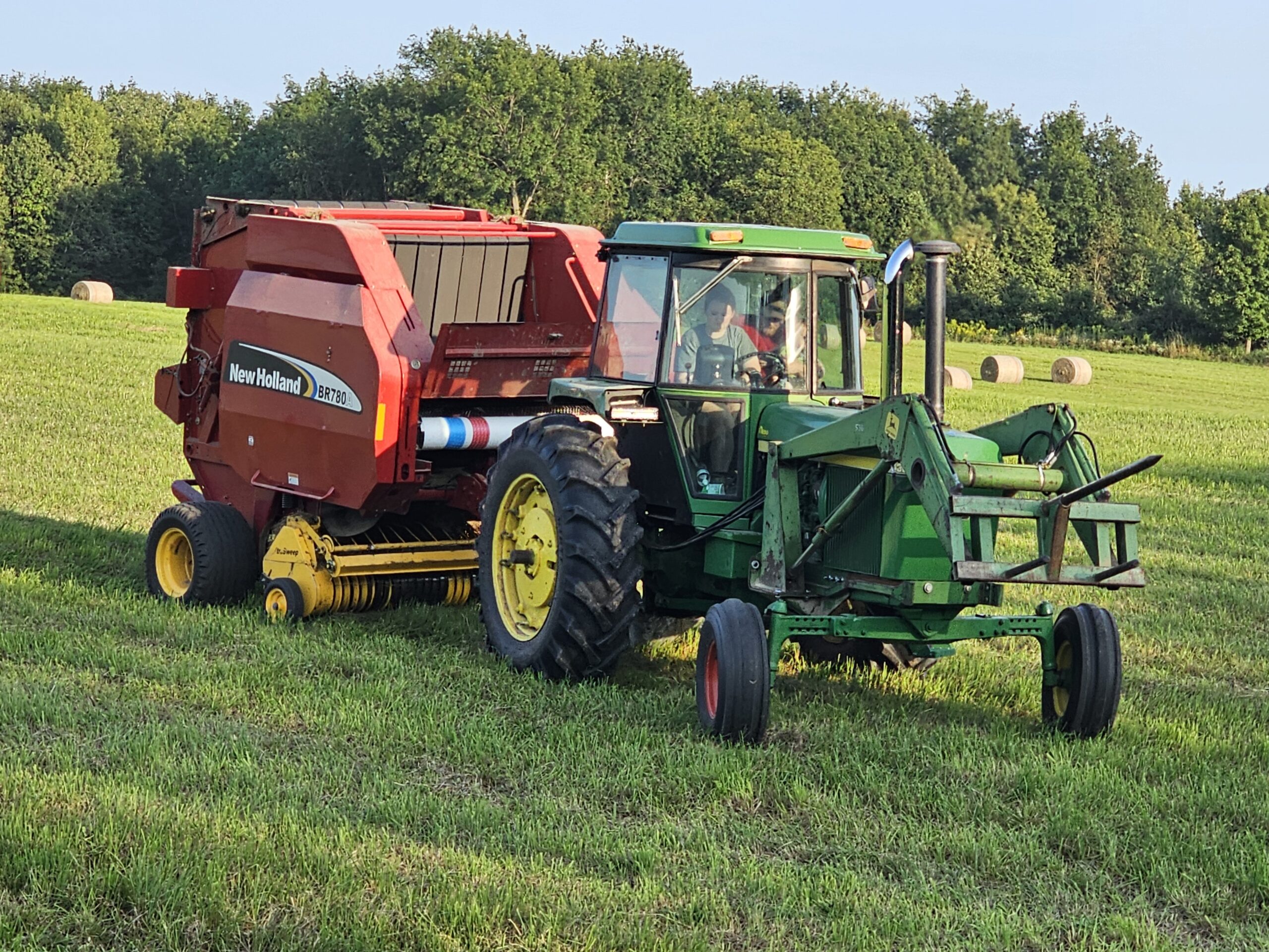Green tractor with loader attachment operating a New Holland BR780 round baler in a grassy field, with hay bales visible in the background.